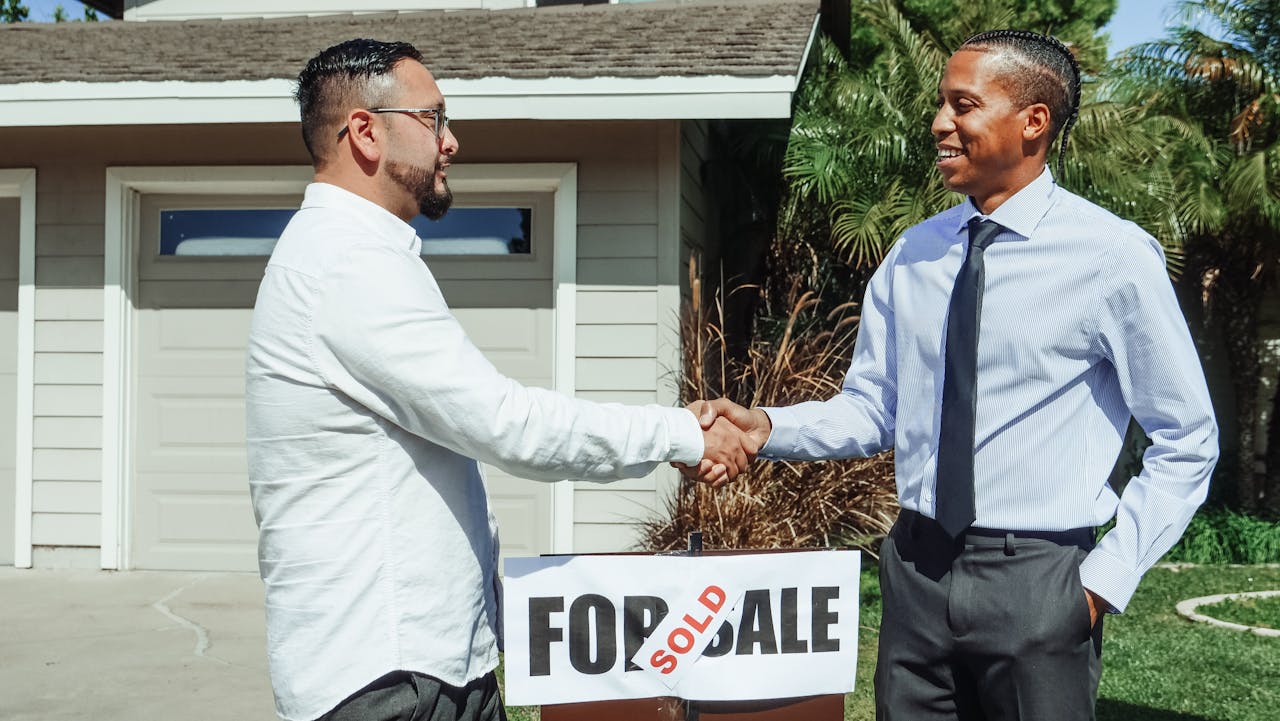 Mastering the First Impression: Your intriguing post title goes here Two men shaking hands in front of house sold sign, sealing real estate deal outdoors.